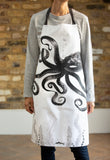 A slim woman wearing an Octopus illustrated white cotton apron with black ties infant of a plain brick wall. A black and grey loose brush drawing of a floating octopus