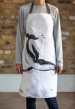 A slim woman wearing a Whale illustrated white cotton apron with black ties infant of a plain brick wall. A large black and grey loose brush drawing of a diving humpback whale