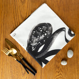Folded Octopus napkin into a square on an oak table next to gold cutlery and two tiny pebbles
